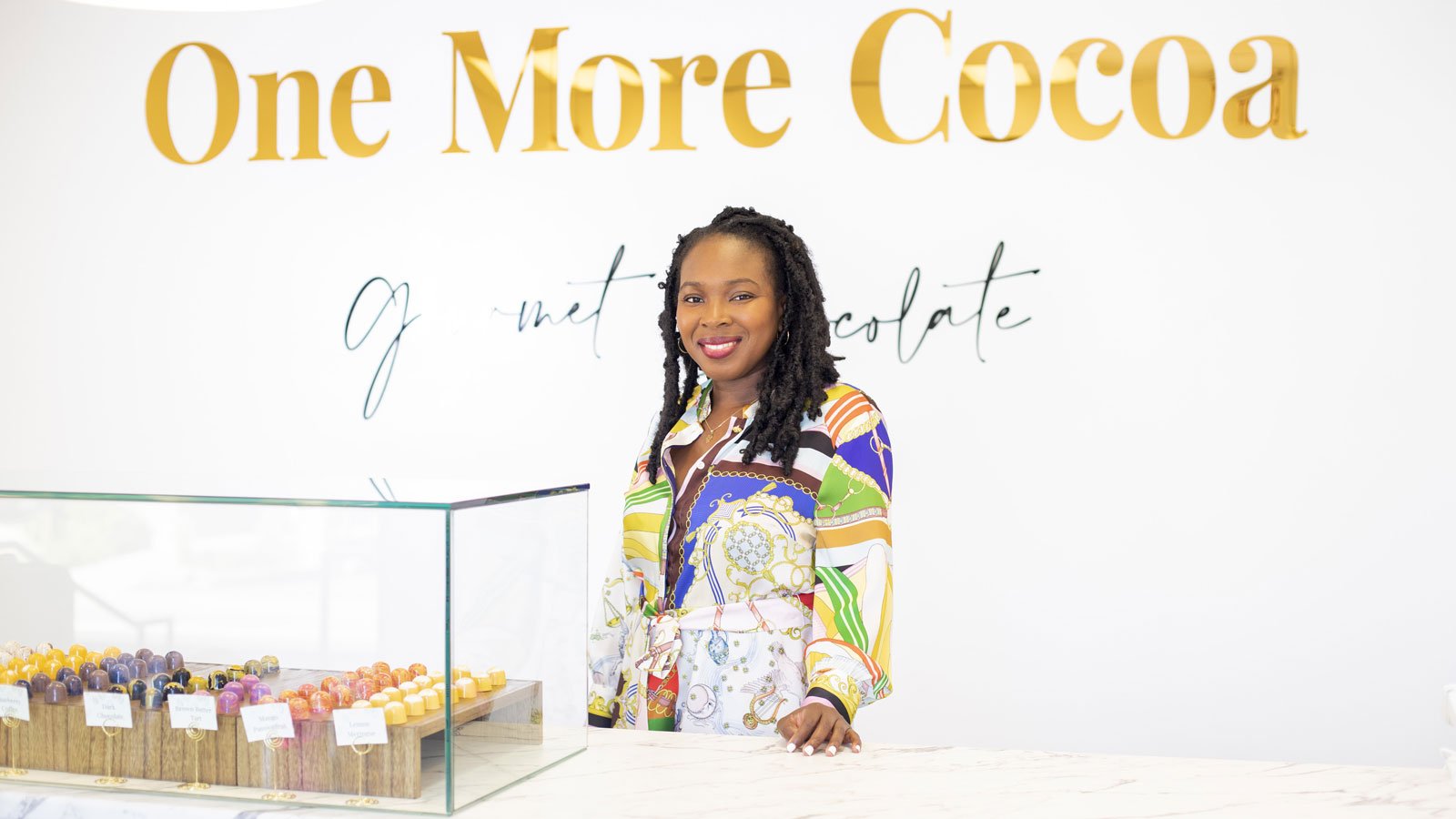 Owner Kenesha Lewis smiling and standing behind a glass display case of chocolate bonbons”