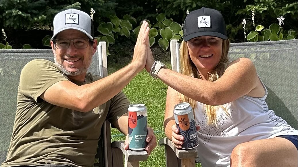 5 Paddles Brewing owners Tristian and Sarah sitting outdoors, both holding a canned beer and high-fiving