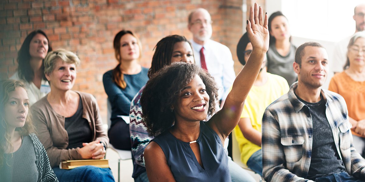 A group of community members engage in a Town-Hall-style discussion, with a woman raising her hand to speak.