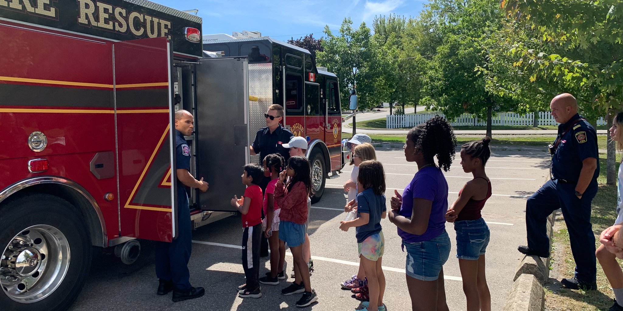 A group of campers look on as firefighters display different features of a red fire truck.